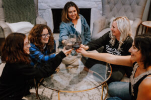 five caucasian women hold glasses of wine together over a glass coffee table in a sitting room