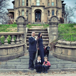 a family dressed as Addams family characters in front of a stone banister entrance to a large house