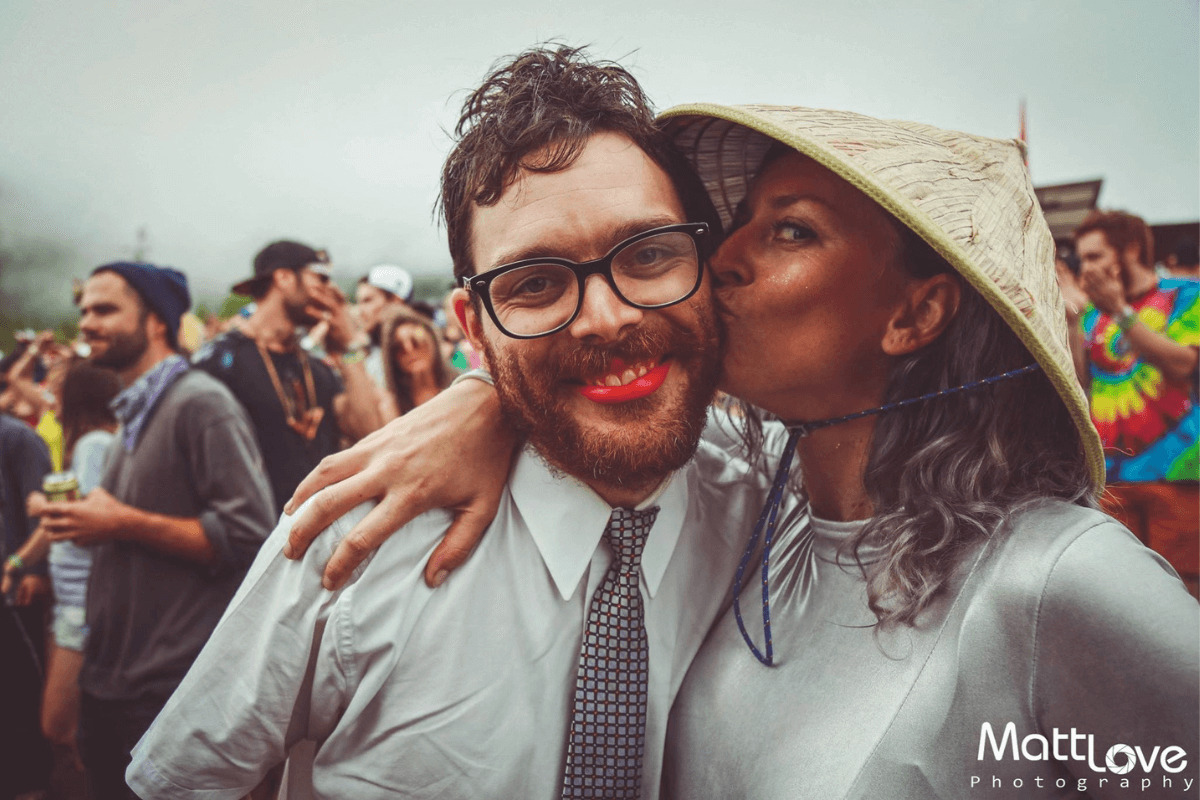 a woman in a hat kisses a man's cheek as he smiles at the camera