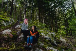 two caucasian women sit on rocks in a forest, smiling at each other