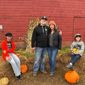a family of four posing in front of hay bales, pumpkins, and the side of red barn
